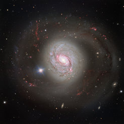 Eine farbenreiche Aufnahme der Spiralgalaxie Messier 77 (M77), auch bekannt als NGC 1068. Die Galaxie zeigt eine beeindruckende Struktur mit hell leuchtendem Zentrum und spiralförmig angeordneten Armen, die sich weit ins All erstrecken. Die Spiralarme sind durch rosafarbene und rote Regionen hervorgehoben, welche Gebiete aktiver Sternentstehung darstellen. Umgeben wird die Galaxie von unzähligen kleinen Lichtpunkten, die ferne Sterne und Galaxien darstellen. Im Vordergrund leuchtet ein heller, sternähnlicher Punkt auf der linken Seite der Galaxie. Die Farben der Galaxie reichen von weiß und blau im Zentrum bis hin zu rötlichen Tönen in den äußeren Bereichen.
